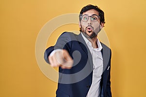 Handsome latin man standing over yellow background pointing with finger surprised ahead, open mouth amazed expression, something