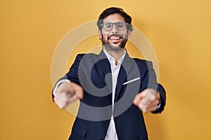 Handsome latin man standing over yellow background pointing to you and the camera with fingers, smiling positive and cheerful