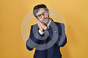 Handsome latin man standing over yellow background sleeping tired dreaming and posing with hands together while smiling with