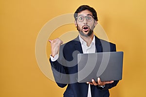 Handsome latin man working using computer laptop surprised pointing with hand finger to the side, open mouth amazed expression