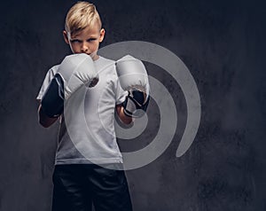 A handsome little boy boxer with blonde hair dressed in a white t-shirt in gloves ready to fight. Isolated on a dark