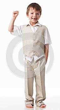 A handsome little boy dressed as a schoolboy - in a white shirt and gray trousers, stands against a white wall and poses