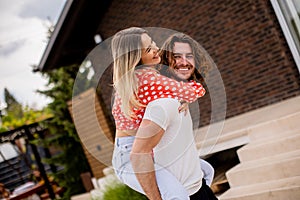 Handsome long hair man carrying the young woman on his back in front of brick house