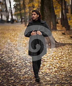 Handsome long haired man in black trench coat walking in the park in wonderful autumn day
