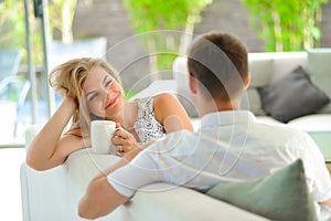 Handsome long haired young blonde woman sitting on a couch propped her head with one hand and holding a mug of delicious tea in an