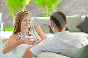 Handsome long haired young blonde woman sitting on a couch propped her head with one hand and holding a mug of delicious tea in an
