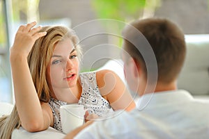 Handsome long haired young blonde woman sitting on a couch propped her head with one hand and holding a mug of delicious tea in an