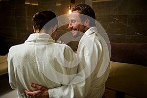 Handsome loving Caucasian man hugging his beloved wife, sitting close to each other while resting together at wellness spa resort