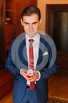 Handsome luxury dressed man in stylish blue holding red heart-shaped box with wedding rings. Classic wooden room interior as