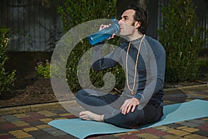 Handsome male athlete, a man yogi with rosary beads, drinking water during yoga practice on a fitness mat at sunset.