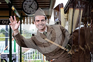 Handsome male British soldier in WW2 vintage uniform at train station leaning out of train, waving and smiling