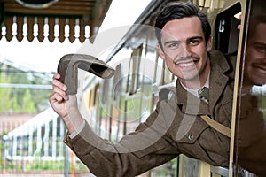 Handsome male British soldier in WW2 vintage uniform at train station leaning out of train window, waving, smiling