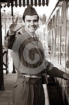Handsome male British soldier in WW2 vintage uniform at train station next to train, waving and smiling