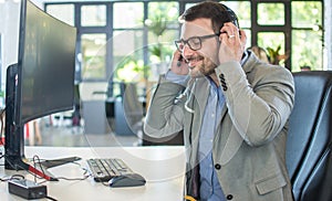 Handsome male call center operator in headset working with computer in modern office, putting on headset over his head.