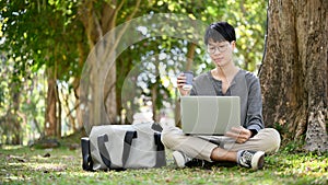 Handsome male college student wearing glasses, sipping coffee while using laptop in the park