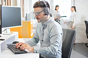 Handsome male customer support phone operator with headset working on desktop computer in office