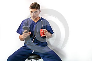 Handsome male doctor in blue scrubs, sitting on stool using phone while drinking from mug