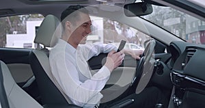 Handsome male entrepreneur sits in the driver's seat in a car and uses a modern smartphone. Smiling man businessman in