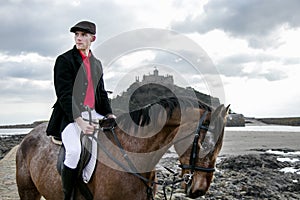 Handsome Male Horse Rider riding horse on beach in traditional fashion with St Michael`s Mount in background