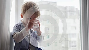 Handsome male kid holds glass cup with clear clear drinking cool water and removes thirst while sitting by window