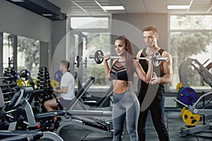 Male personal trainer supporting woman working with heavy barbell at gym.