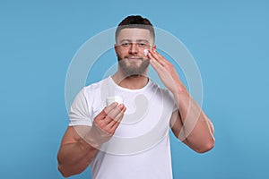 Handsome man applying cream onto his face on light blue background