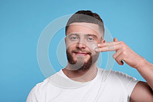 Handsome man applying cream onto his face on light blue background