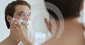 Handsome man applying shaving foam on face looking in mirror