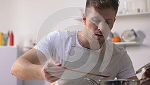 Handsome man in apron cooking, stirring ingredients in pan and trying meal