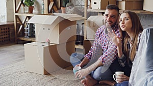 Handsome man and attractive woman are sitting on floor in new house and planning interior of their new home holding mugs