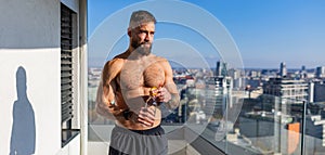 Handsome man with bare chest standing on balcony and enjoying beautiful view on city. Concept of selfcare and enjoying