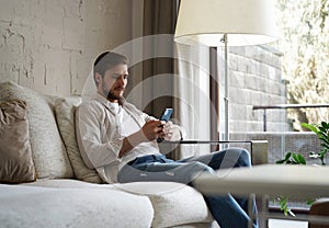 Handsome man in basic t-shirt smiling and holding mobile phone in hands while sitting on couch in living room.