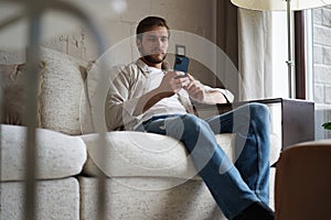 Handsome man in basic t-shirt smiling and holding mobile phone in hands while sitting on couch in living room.