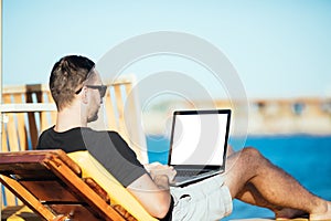 Handsome man on the beach typing on his laptop with white screen for copyspace while sitting on his deck chair