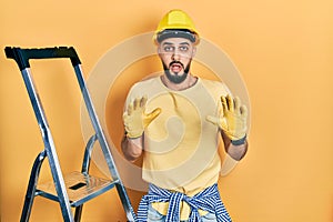 Handsome man with beard by construction stairs wearing hardhat afraid and terrified with fear expression stop gesture with hands,
