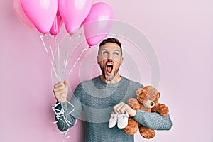 Handsome man with beard expecting a baby girl holding balloons, shoes and teddy bear angry and mad screaming frustrated and