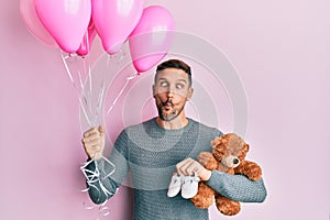 Handsome man with beard expecting a baby girl holding balloons, shoes and teddy bear making fish face with mouth and squinting