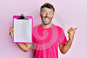 Handsome man with beard holding clipboard showing blank space pointing thumb up to the side smiling happy with open mouth