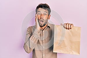 Handsome man with beard holding delivery paper bag with heart reminder afraid and shocked, surprise and amazed expression with