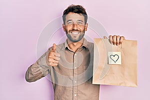 Handsome man with beard holding delivery paper bag with heart reminder doing happy thumbs up gesture with hand