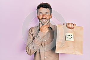 Handsome man with beard holding delivery paper bag with heart reminder looking confident at the camera smiling with crossed arms