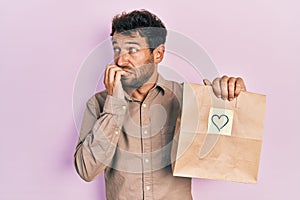 Handsome man with beard holding delivery paper bag with heart reminder looking stressed and nervous with hands on mouth biting