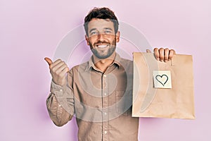 Handsome man with beard holding delivery paper bag with heart reminder pointing to the back behind with hand and thumbs up,