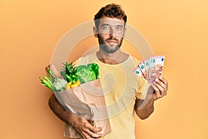 Handsome man with beard holding groceries and 10 euros banknotes relaxed with serious expression on face