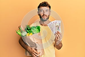 Handsome man with beard holding groceries and 100 ruble banknotes relaxed with serious expression on face