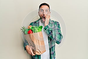 Handsome man with beard holding paper bag with groceries afraid and shocked, surprise and amazed expression with hands on face