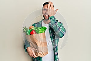 Handsome man with beard holding paper bag with groceries covering one eye with hand, confident smile on face and surprise emotion