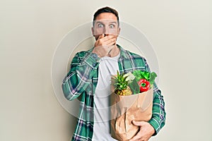 Handsome man with beard holding paper bag with groceries shocked covering mouth with hands for mistake