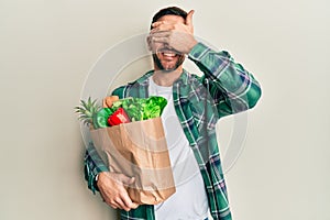 Handsome man with beard holding paper bag with groceries smiling and laughing with hand on face covering eyes for surprise