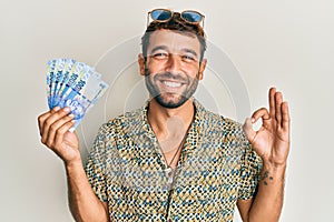 Handsome man with beard holding south african 100 rand banknotes doing ok sign with fingers, smiling friendly gesturing excellent